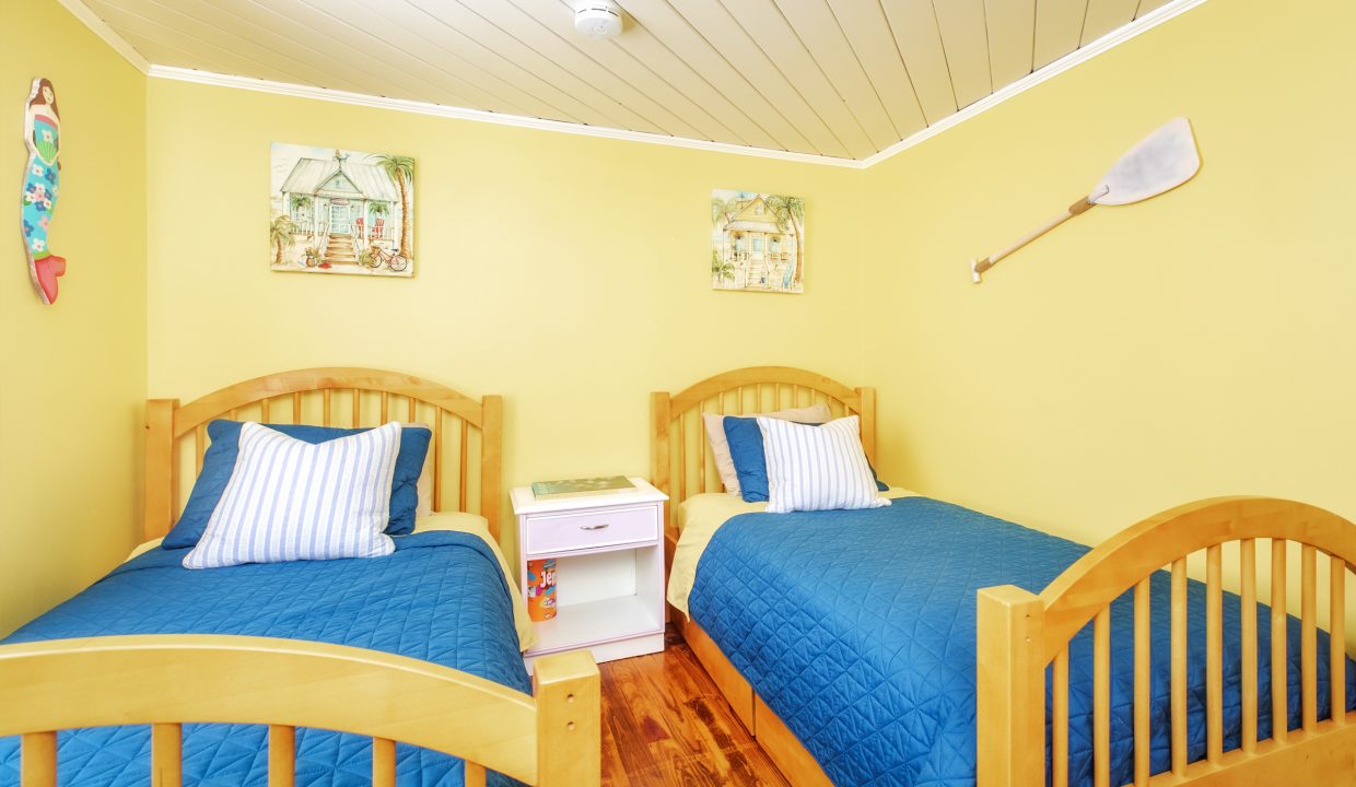 A bedroom with two single wooden beds, blue bedding, a white nightstand between them, yellow walls, nautical-themed decor, and wood flooring.