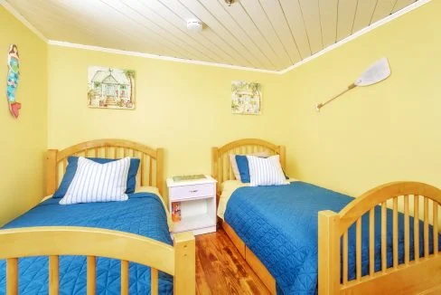 A bedroom with two single wooden beds, blue bedding, a white nightstand between them, yellow walls, nautical-themed decor, and wood flooring.