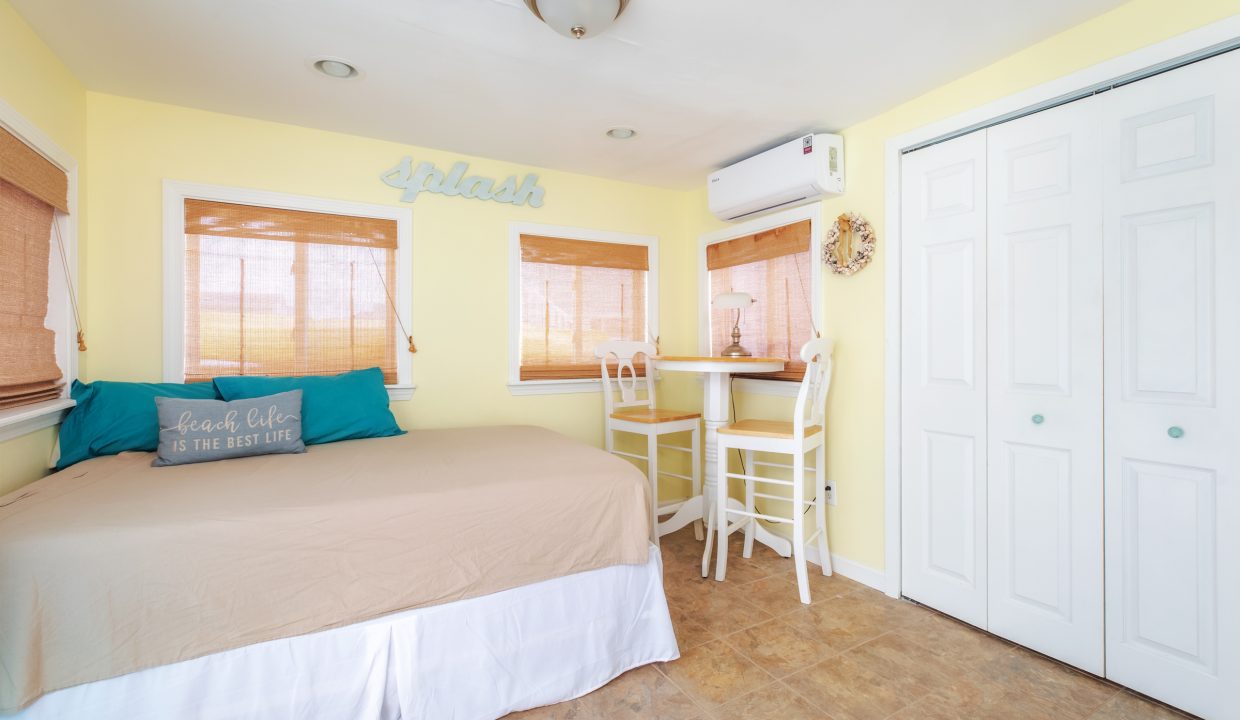 A small room features a bed, a wall-mounted air conditioner, and a round table with two chairs. The walls are painted yellow, and there are three windows with shades. A closet with white doors is on the right.