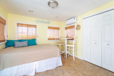 A small room features a bed, a wall-mounted air conditioner, and a round table with two chairs. The walls are painted yellow, and there are three windows with shades. A closet with white doors is on the right.