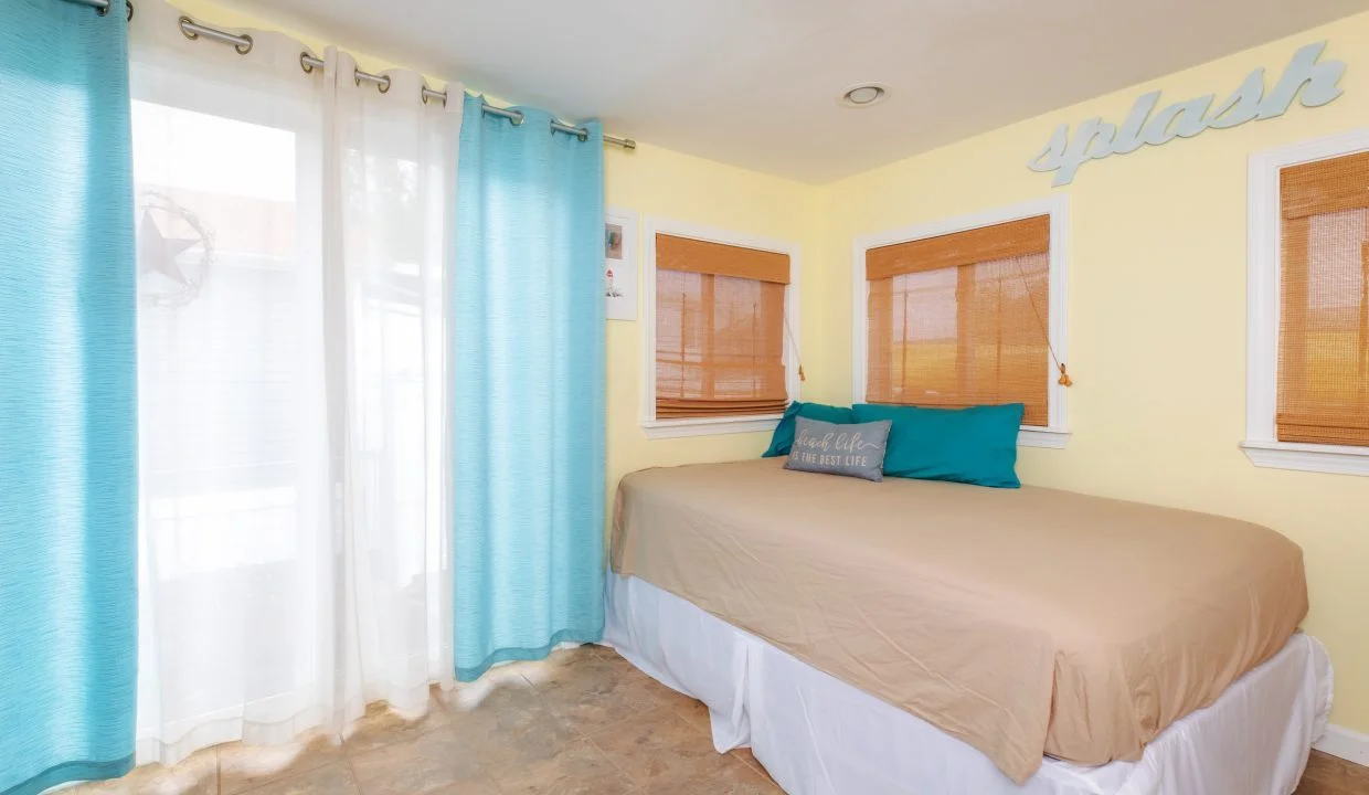 A small bedroom with a tan bed, teal and beige pillows, yellow walls, windows with brown blinds, turquoise curtains, and the word 