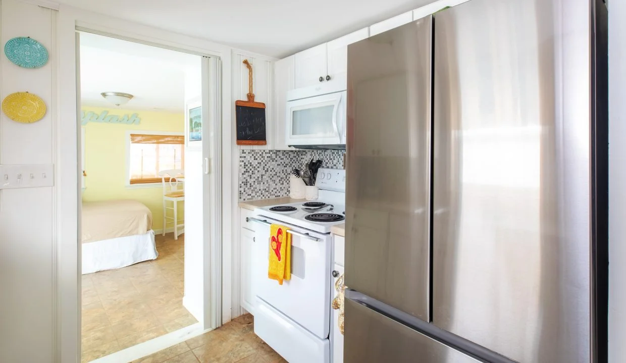 A compact kitchen with white cabinets, stainless steel refrigerator, electric stove, and microwave. A glimpse of a bedroom with a bed and chair is visible through an adjoining door.