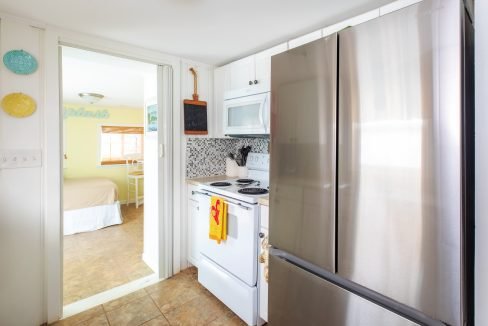 A compact kitchen with white cabinets, stainless steel refrigerator, electric stove, and microwave. A glimpse of a bedroom with a bed and chair is visible through an adjoining door.