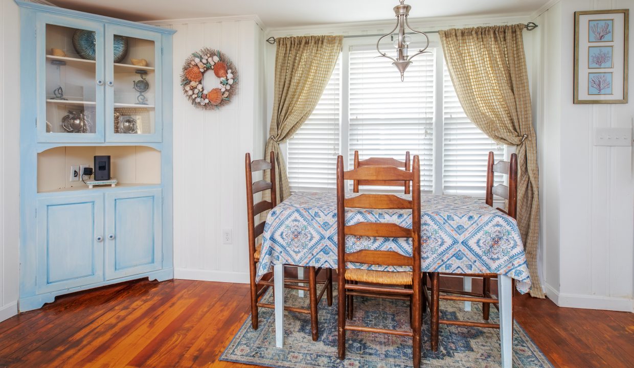 A dining room with a wooden table covered with a patterned tablecloth, six chairs, a light fixture, and a blue cabinet. There are two windows with patterned curtains and a wall wreath.