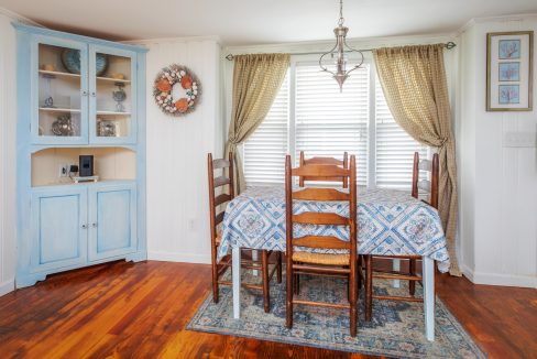 A dining room with a wooden table covered with a patterned tablecloth, six chairs, a light fixture, and a blue cabinet. There are two windows with patterned curtains and a wall wreath.