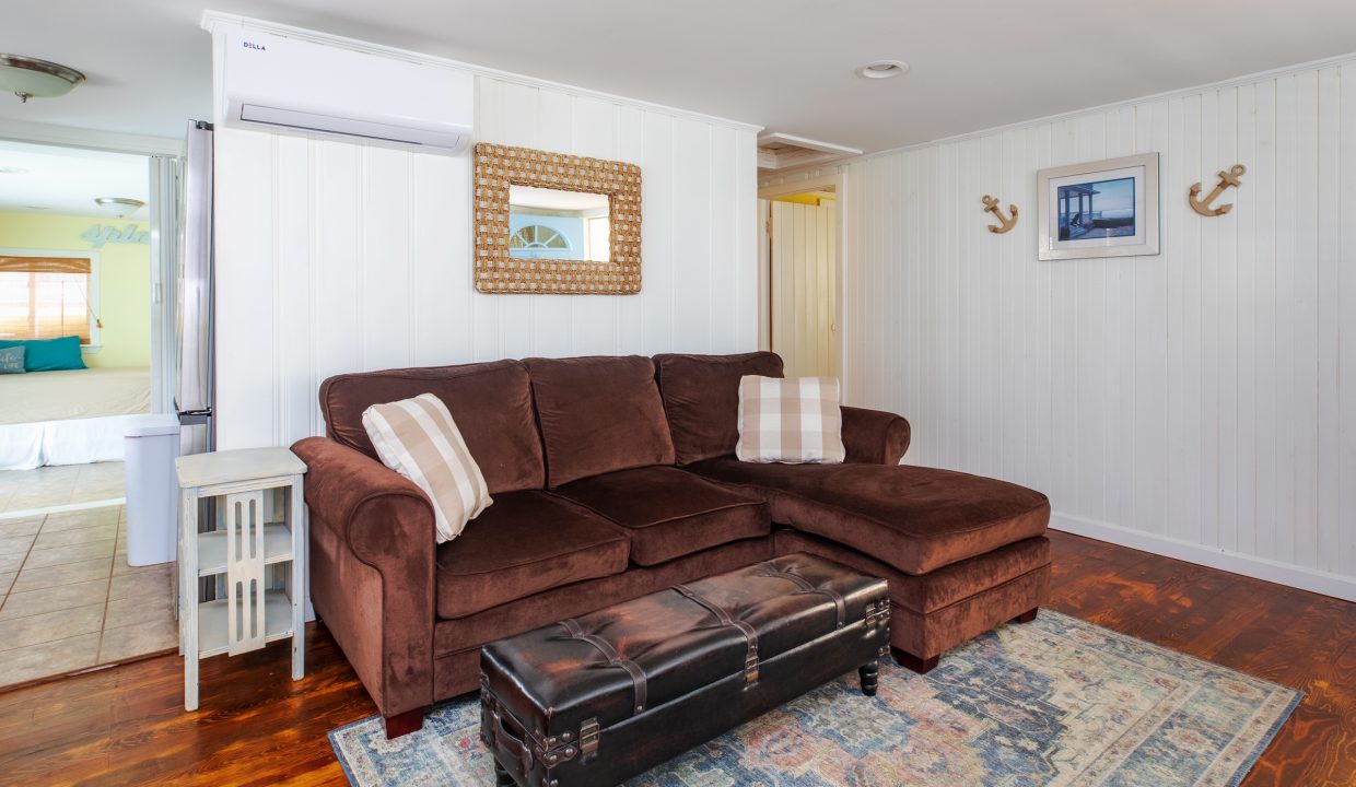 A cozy living room features a brown sectional sofa with throw pillows, a decorative mirror, wall art, and a patterned rug. An adjacent doorway leads to a bedroom with a window.