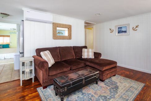 A cozy living room features a brown sectional sofa with throw pillows, a decorative mirror, wall art, and a patterned rug. An adjacent doorway leads to a bedroom with a window.