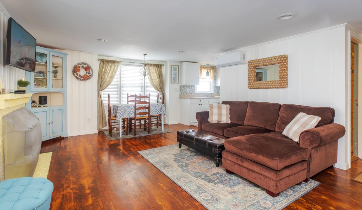 A cozy living room with a brown sectional sofa, a television mounted on the wall, and a fireplace. The dining area is visible with a table and chairs near a window in the background.