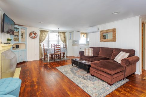 A cozy living room with a brown sectional sofa, a television mounted on the wall, and a fireplace. The dining area is visible with a table and chairs near a window in the background.