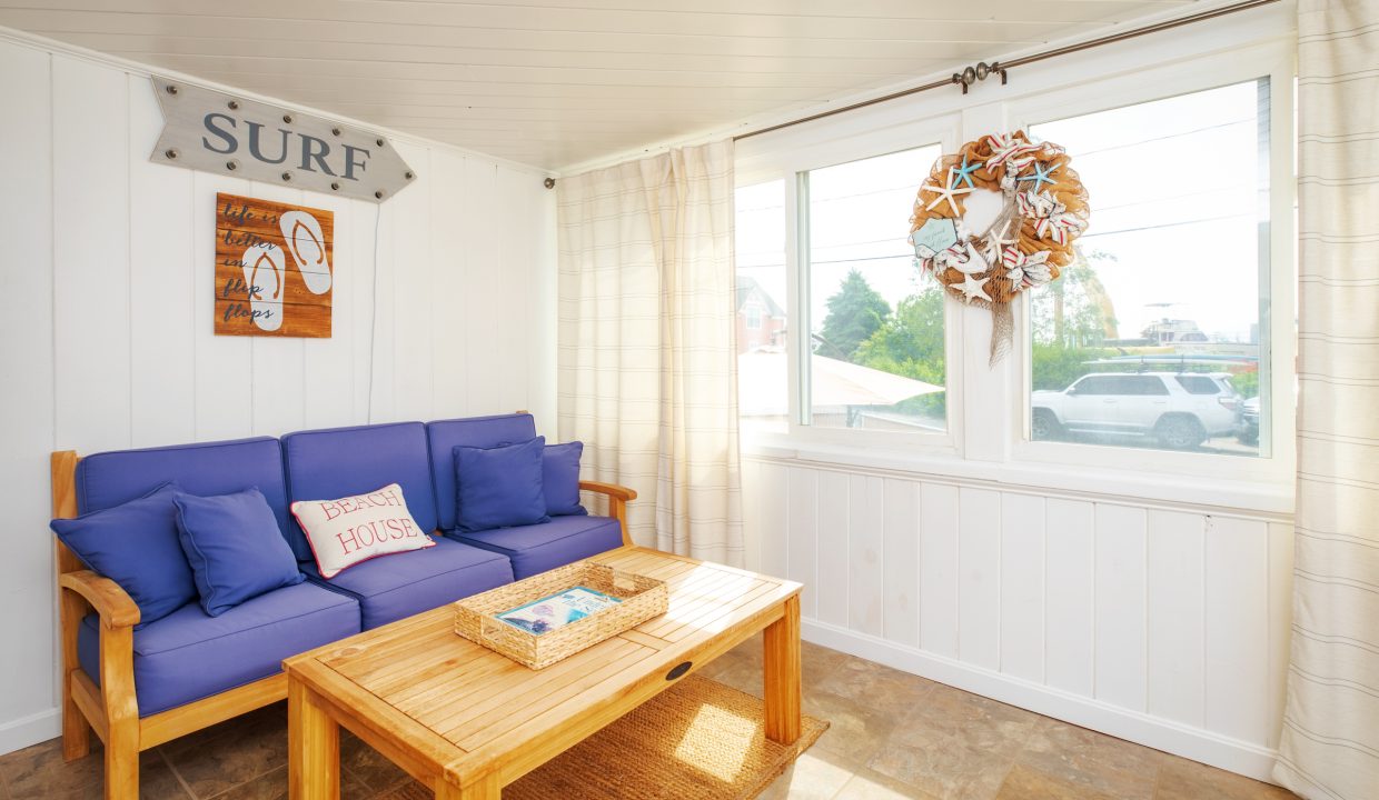 Bright sunroom with white walls, blue cushioned wooden sofa, decorative surf sign, wreath on the window, and wooden coffee table with a woven tray. Large windows offer a view of the outdoors.