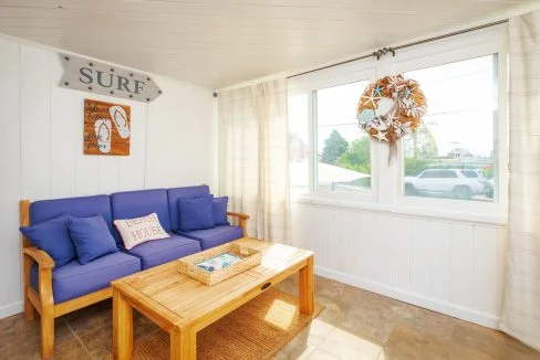 Bright sunroom with white walls, blue cushioned wooden sofa, decorative surf sign, wreath on the window, and wooden coffee table with a woven tray. Large windows offer a view of the outdoors.