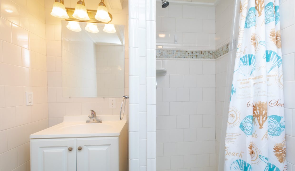 A bathroom with white tile walls, a sink with a mirror under three lights, and a shower with a curtain decorated with seashell designs.