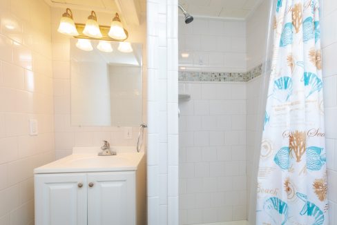 A bathroom with white tile walls, a sink with a mirror under three lights, and a shower with a curtain decorated with seashell designs.