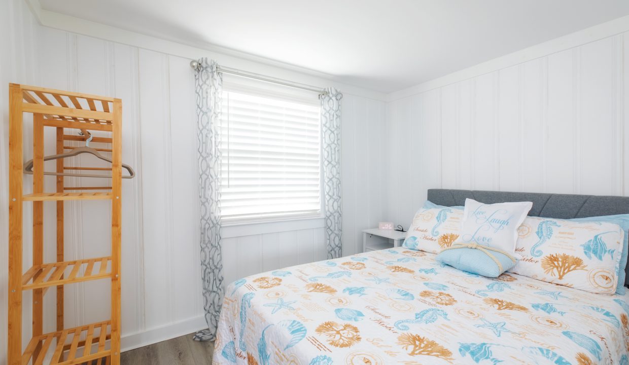 A bedroom with white paneled walls, a window with patterned curtains, a wooden shelf, and a bed with a beach-themed quilt and decorative pillows.