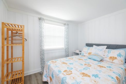 A bedroom with white paneled walls, a window with patterned curtains, a wooden shelf, and a bed with a beach-themed quilt and decorative pillows.