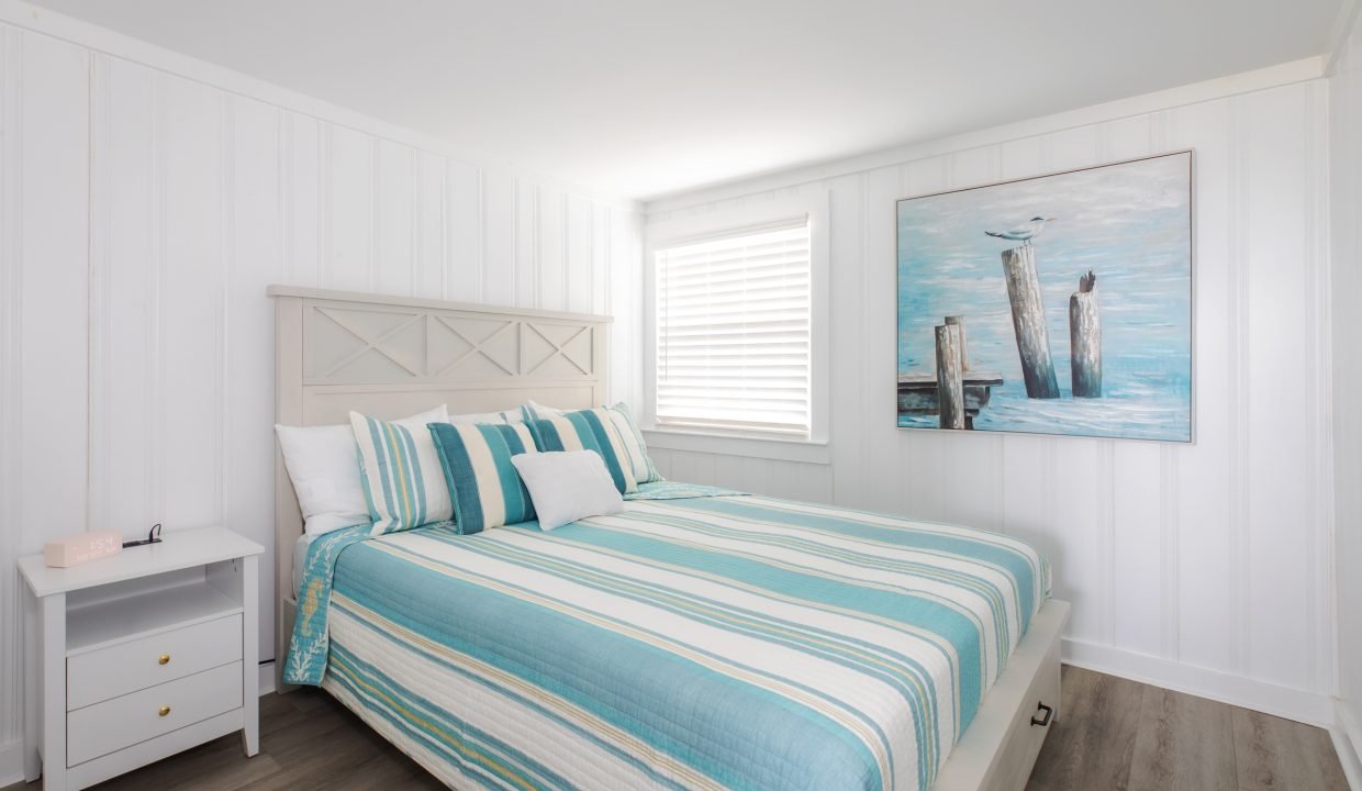 A bedroom with white panel walls, a bed with blue and white striped bedding, a nightstand with a pink clock, and a painting of seagulls on wooden posts above the bed.