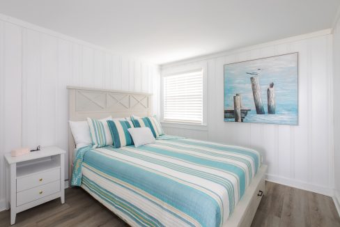 A bedroom with white panel walls, a bed with blue and white striped bedding, a nightstand with a pink clock, and a painting of seagulls on wooden posts above the bed.