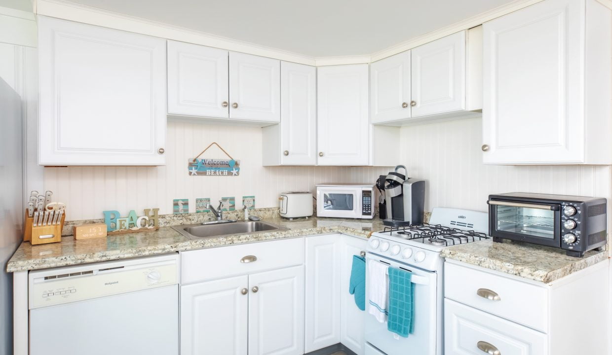 A small kitchen with white cabinets, a marble countertop, a stove, a microwave, a toaster oven, a dishwasher, and various kitchen utensils. A sign above the sink says, 