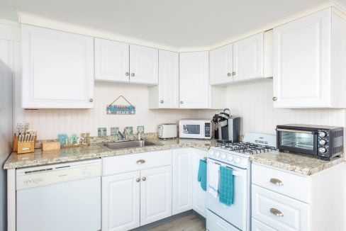 A small kitchen with white cabinets, a marble countertop, a stove, a microwave, a toaster oven, a dishwasher, and various kitchen utensils. A sign above the sink says, 