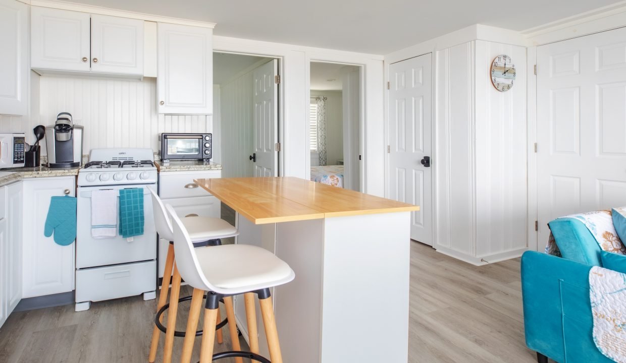 A bright, modern kitchen with white cabinets, a wooden island with stools, a stove, microwave, coffee maker, and teal accents. Adjacent rooms and a wall clock are visible in the background.