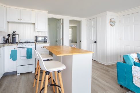 A bright, modern kitchen with white cabinets, a wooden island with stools, a stove, microwave, coffee maker, and teal accents. Adjacent rooms and a wall clock are visible in the background.