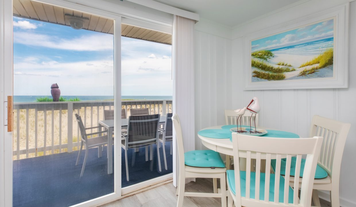 A bright room with a small round dining table and chairs, large sliding glass doors leading to a balcony with a beach view, and a coastal-themed painting on the wall.