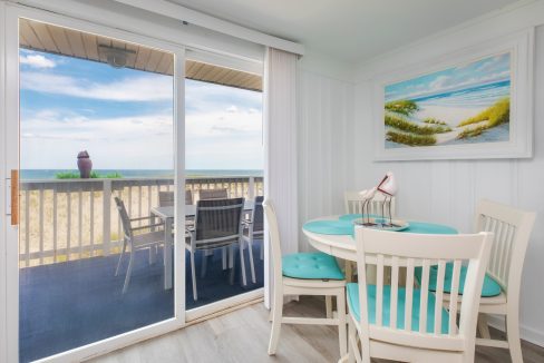 A bright room with a small round dining table and chairs, large sliding glass doors leading to a balcony with a beach view, and a coastal-themed painting on the wall.