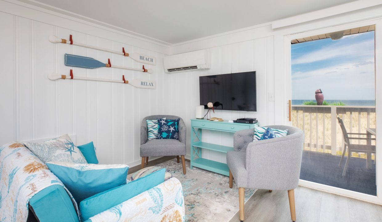 A cozy living room with a beach-themed decor, featuring white walls, a couch, two armchairs, a television, and a window offering a view of the ocean with a balcony area outside.