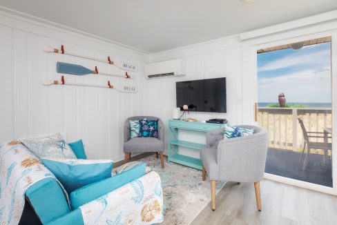 A cozy living room with a beach-themed decor, featuring white walls, a couch, two armchairs, a television, and a window offering a view of the ocean with a balcony area outside.