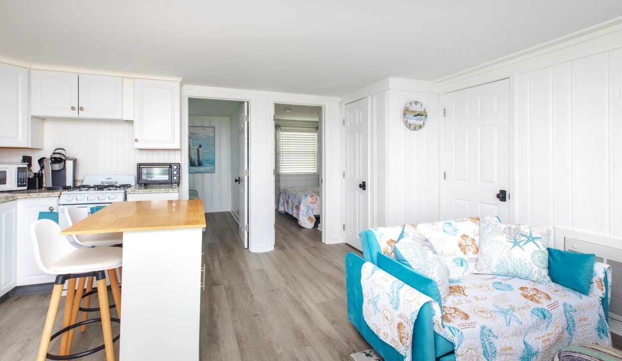 A bright and airy kitchen and living area with white cabinets, wood flooring, a small island with stools, and a turquoise-accented sofa. A doorway leads to a bedroom in the background.