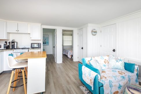 A bright and airy kitchen and living area with white cabinets, wood flooring, a small island with stools, and a turquoise-accented sofa. A doorway leads to a bedroom in the background.