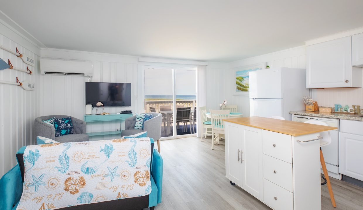 A bright, open-plan living space with a sofa, TV, kitchen area, dining table, and large sliding doors leading to a balcony with an ocean view. The decor includes coastal-themed elements.