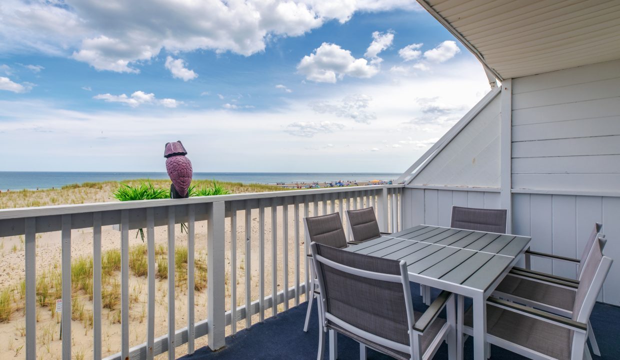 A beachside balcony features a table with four chairs, white railing, and a view of the sandy beach and ocean under a partly cloudy sky. An owl statue sits on the railing.