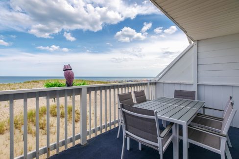 A beachside balcony features a table with four chairs, white railing, and a view of the sandy beach and ocean under a partly cloudy sky. An owl statue sits on the railing.