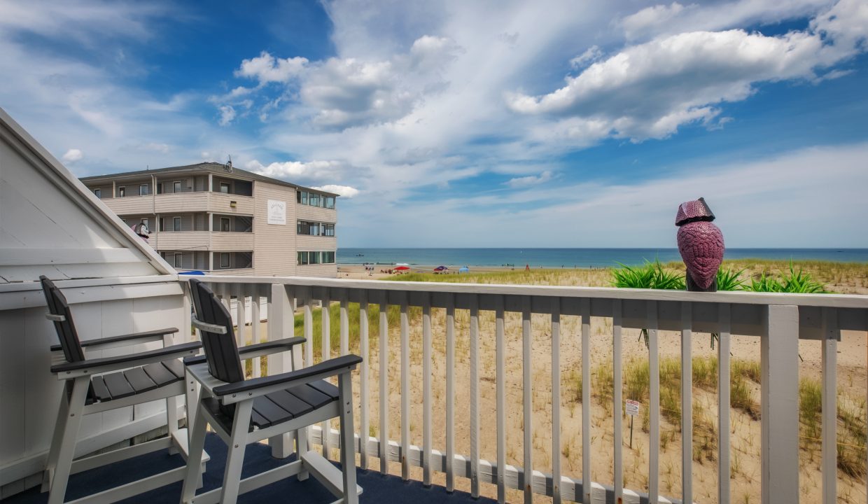 A beachfront balcony with two chairs overlooks the sandy shore and ocean under a partly cloudy sky. A building stands in the background, and a decorative owl statue is perched on the railing.