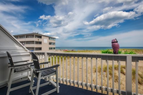 A beachfront balcony with two chairs overlooks the sandy shore and ocean under a partly cloudy sky. A building stands in the background, and a decorative owl statue is perched on the railing.