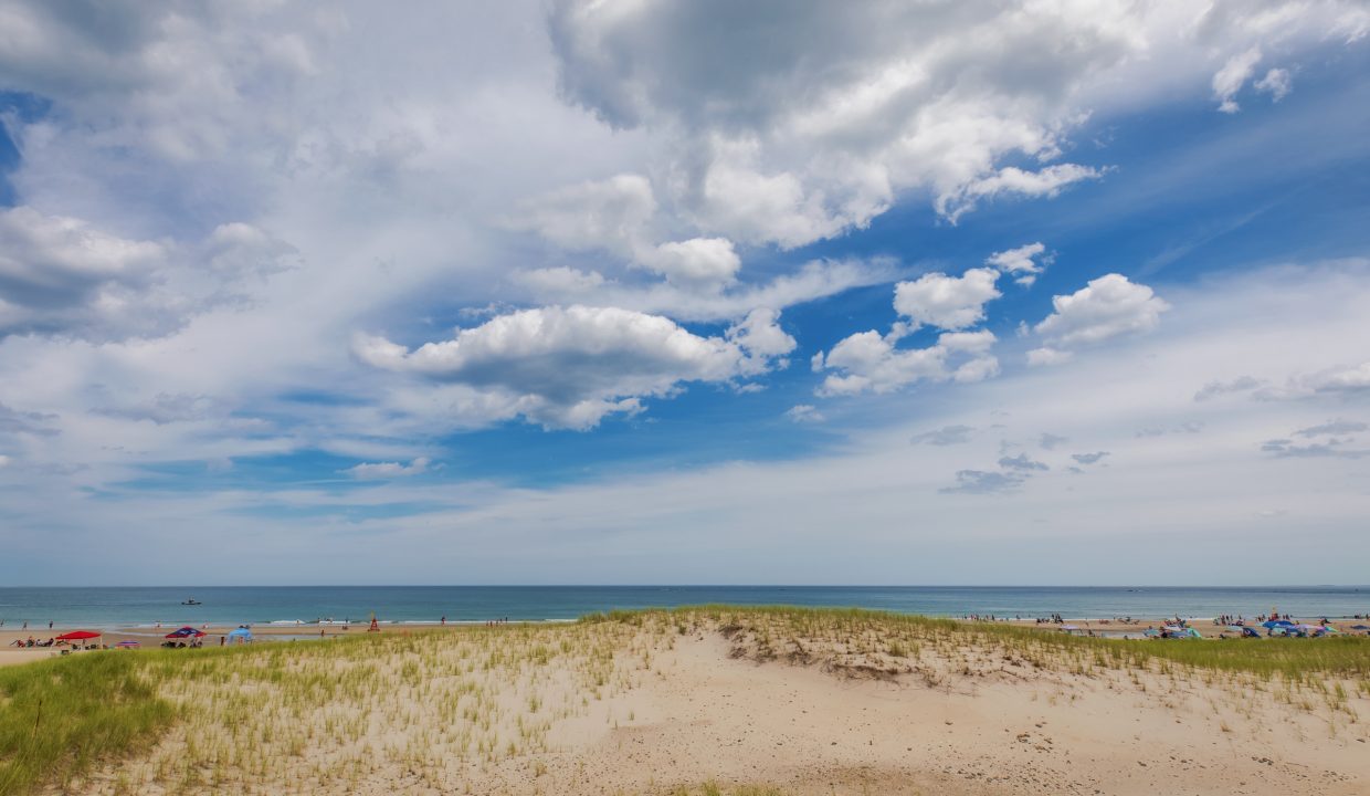 A sandy beach with scattered grass, partially cloudy sky, and people relaxing near the shoreline.