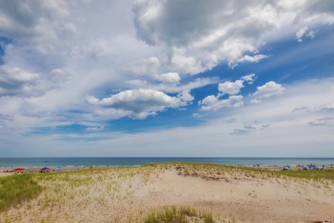 A sandy beach with scattered grass, partially cloudy sky, and people relaxing near the shoreline.