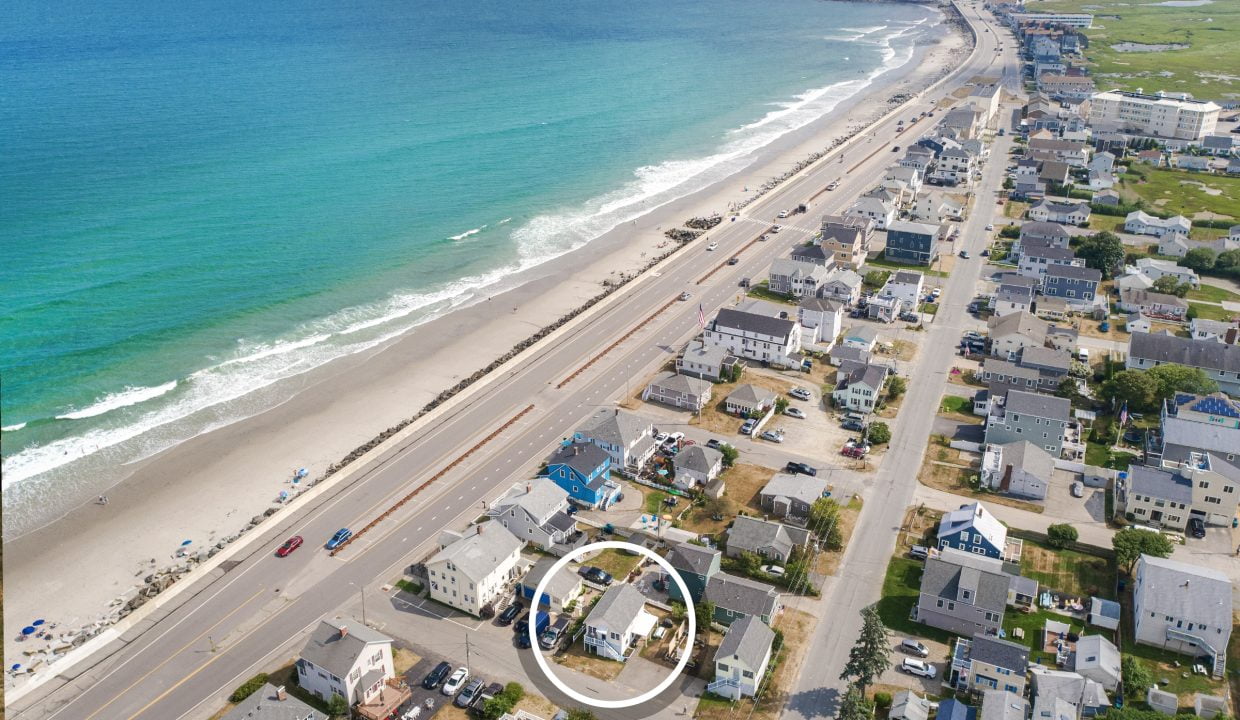 Aerial view of a coastal town featuring streets with houses, a road running parallel to the beach, and a section of the shore with people. A white circle highlights one specific house in the image.