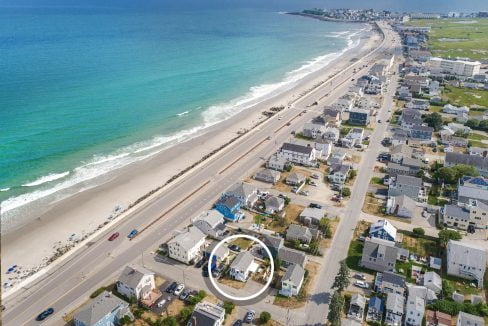 Aerial view of a coastal town featuring streets with houses, a road running parallel to the beach, and a section of the shore with people. A white circle highlights one specific house in the image.