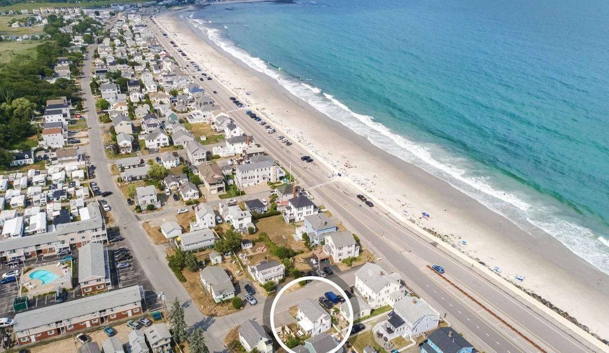 Aerial view of a coastal town with houses lining the shore, a main road running parallel to the beach, and a few people on the sandy shore. A circle highlights a specific property close to the beach.