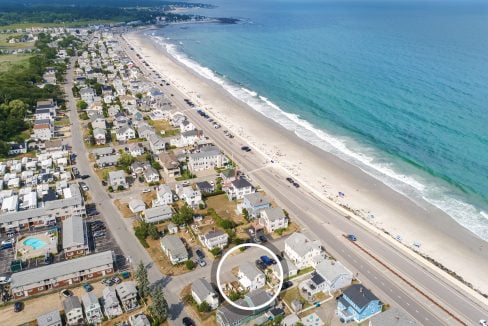 Aerial view of a coastal town with houses lining the shore, a main road running parallel to the beach, and a few people on the sandy shore. A circle highlights a specific property close to the beach.