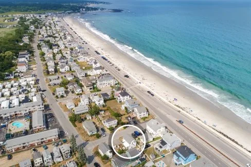 Aerial view of a coastal town with houses lining the shore, a main road running parallel to the beach, and a few people on the sandy shore. A circle highlights a specific property close to the beach.