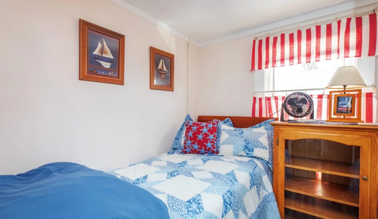 A small bedroom features a bed with blue and white bedding, a wooden nightstand with a fan and lamp, nautical-themed wall art, and windows with red and white striped curtains.