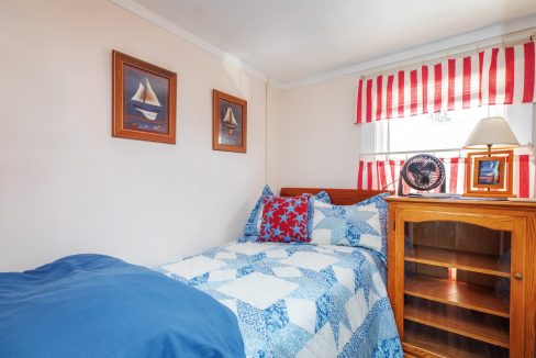 A small bedroom features a bed with blue and white bedding, a wooden nightstand with a fan and lamp, nautical-themed wall art, and windows with red and white striped curtains.