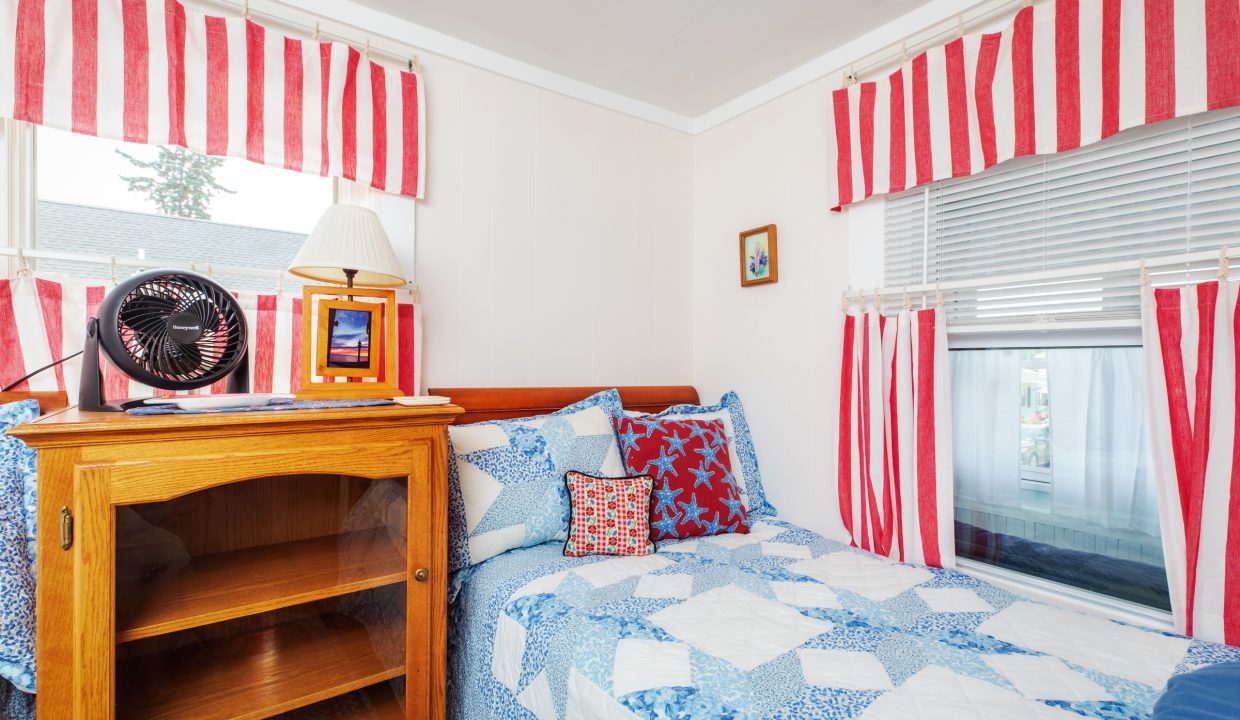 A bedroom featuring a twin bed with a blue and white quilt, a wooden bookshelf, a fan, and decor with red, white, and blue striped patterns on the curtains and pillows.