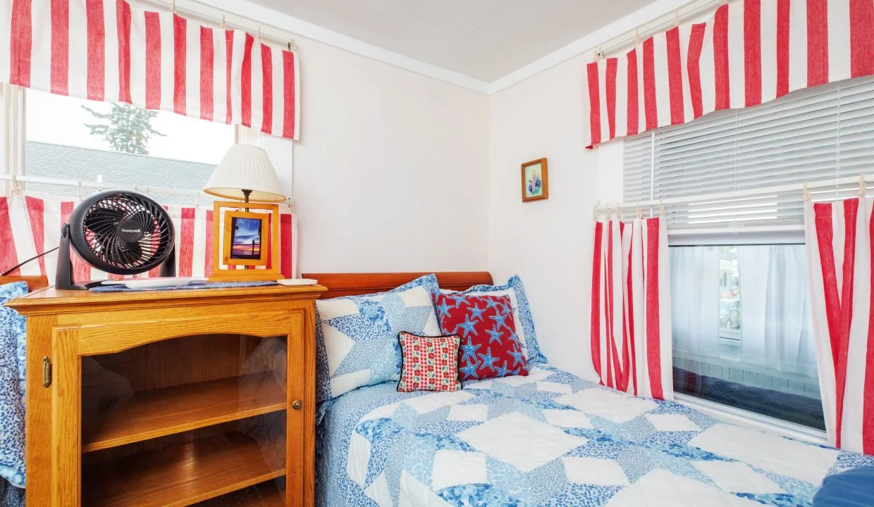 A bedroom featuring a twin bed with a blue and white quilt, a wooden bookshelf, a fan, and decor with red, white, and blue striped patterns on the curtains and pillows.