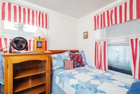 A bedroom featuring a twin bed with a blue and white quilt, a wooden bookshelf, a fan, and decor with red, white, and blue striped patterns on the curtains and pillows.