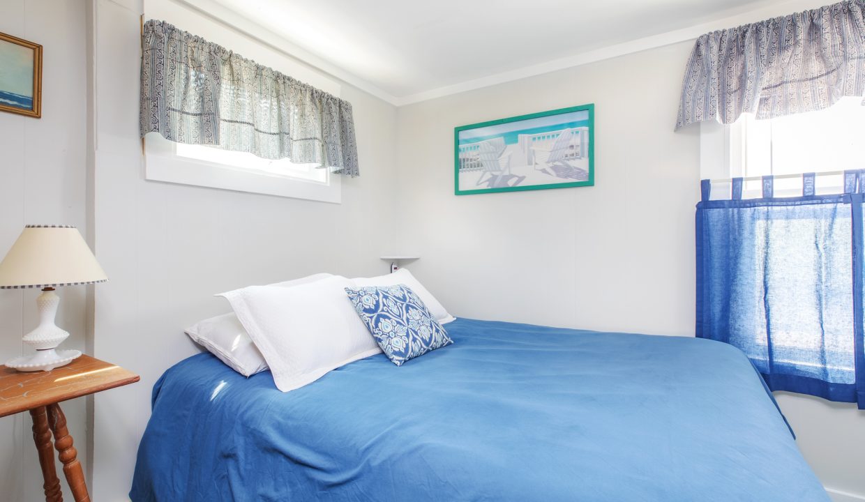 A small bedroom with white walls and a window above the bed. The bed has white pillows, a blue bedspread, and a blue decorative pillow. A nightstand with a lamp is on the left side.