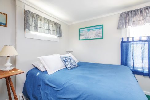 A small bedroom with white walls and a window above the bed. The bed has white pillows, a blue bedspread, and a blue decorative pillow. A nightstand with a lamp is on the left side.